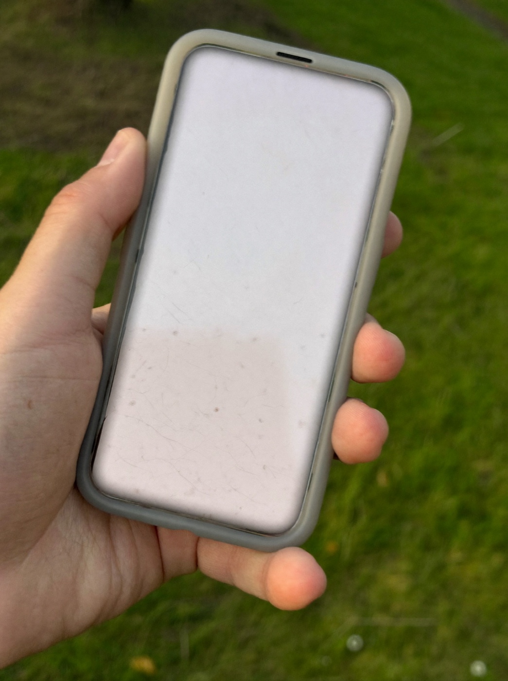 Mão segurando um celular ao ar livre, tela em branco, fundo verde desfocado