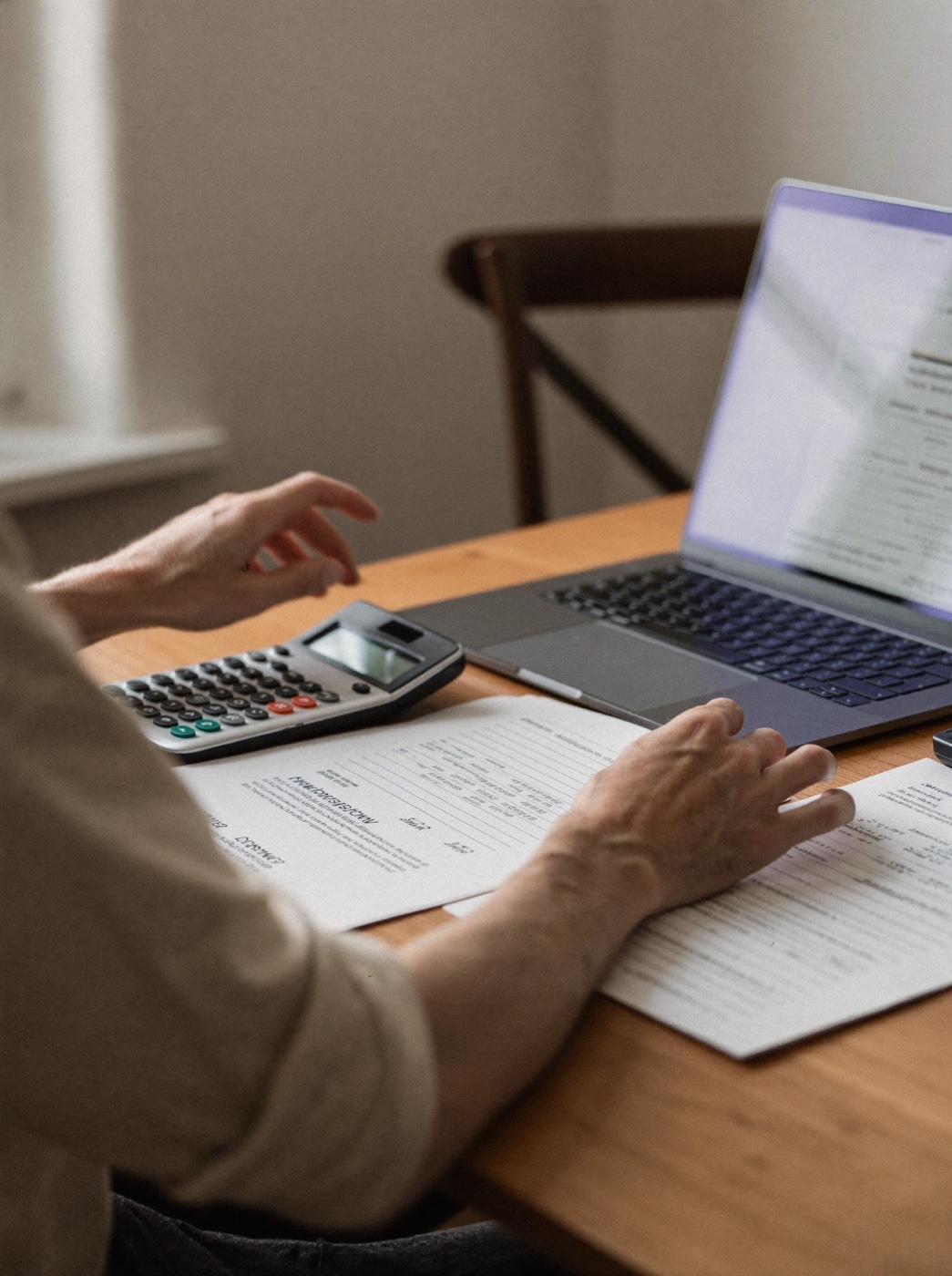 Contador conferindo relatórios impressos com calculadora e laptop numa mesa de madeira