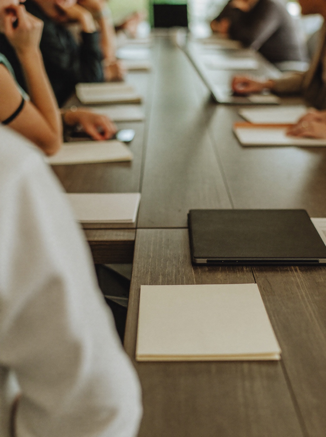 Vários profissionais reunidos ao redor de uma mesa de reunião, vistos de lado e de costas, com notebook fechado e blocos de notas em branco sobre a mesa