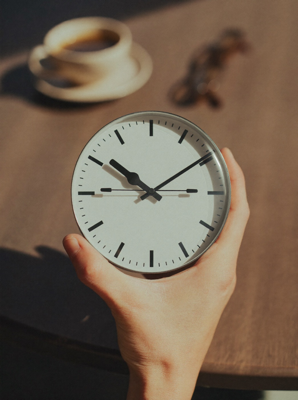 Mão segurando um relógio analógico minimalista sem números, xícara de café ao fundo